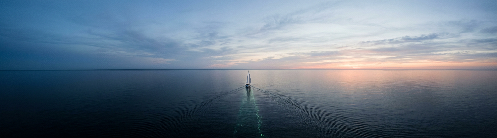 A single sailboat holding a steady course across a calm navy sea toward the horizon at dawn